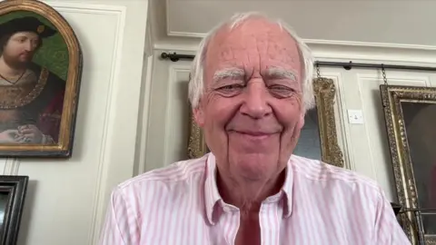 BBC Sir Tim Rice smiles at the camera wearing a pink striped shirt. There are large ornate paintings hung on the wall behind him.