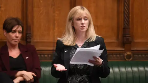 Reuters Rosie Duffield, wearing a dark business suit, stands up to speak in the House of Commons chamber, holding a sheaf of papers in front of her. Her former Labour colleague Jess Phillips sits on the bench behind her.