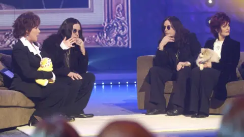Jan Ravens as Sharon Osbourne holds a Pudsey bear toy as she sits alongside Jon Culshaw as Ozzy. The real Ozzy Osbourne sits opposite together with the real Sharon, holding their pet dog. All are dressed in similar black clothing.