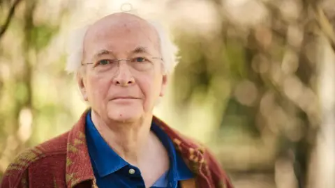 Sam Allard Phillip Pullman, wearing a blue shirt and burgundy jacket