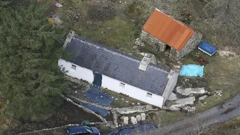 Getty Images An aerial view of a white cottage with a slated roof. There is an old stone outbuilding to its rear with a heavily rusted tin roof. The cottage is beside a tall tree and the front is surrounded by a stone wall. There are a number of black plastic sheets around the property.