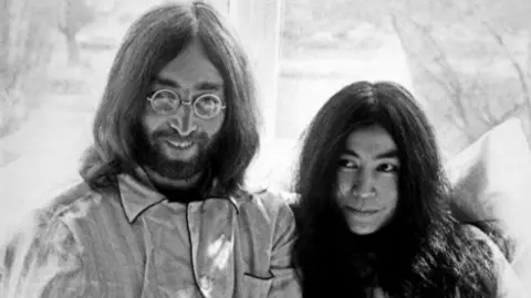 Unknown John Lennon, in round glasses, with long hair and wearing pyjamas, sits on a bed with Yoko Ono as part of their 1969 'Bed-In For Peace' at the Amsterdam Hilton hotel.