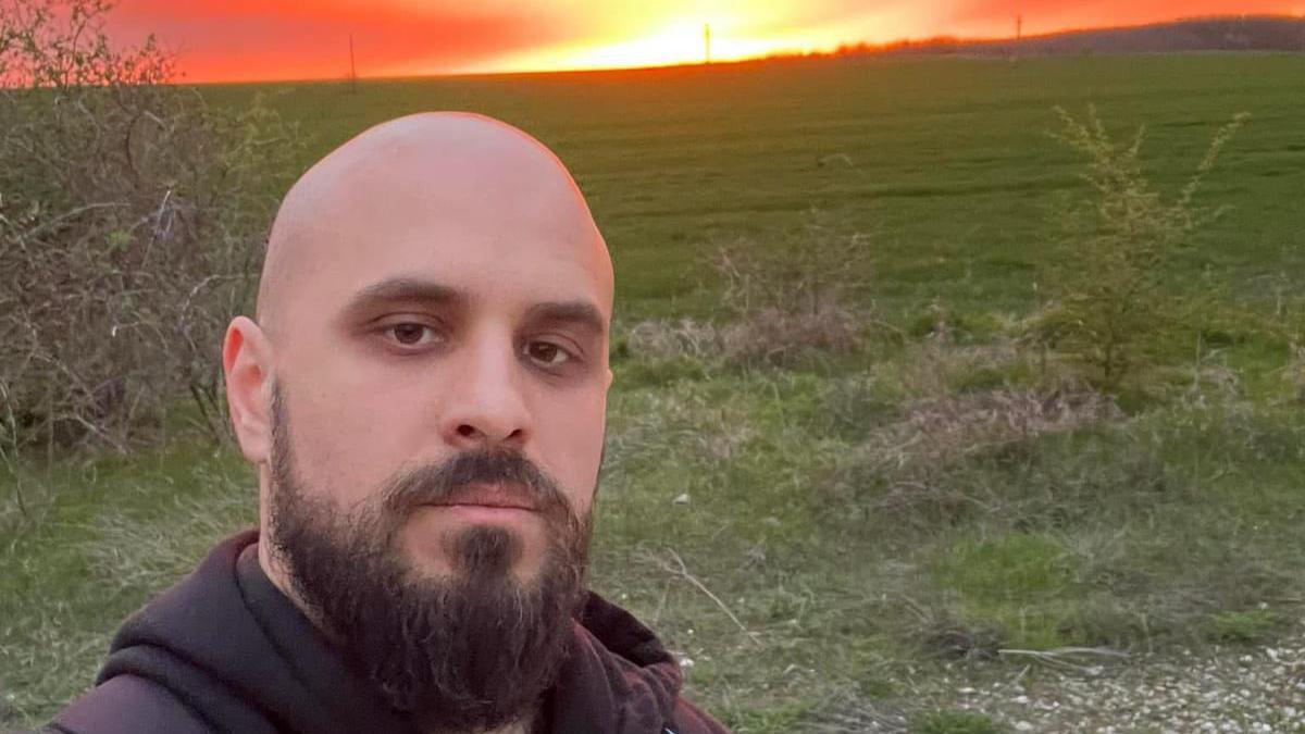 A selfie of a bald white man with a beard, with a sunset behind him