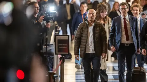 EPA Defendant Yunice Abbas walking in the corridor of the court, next to his lawyer, with a cameraman taking photos from the side