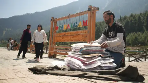 Seraj Ali The image shows a shawl seller sitting next to a tourist board that says