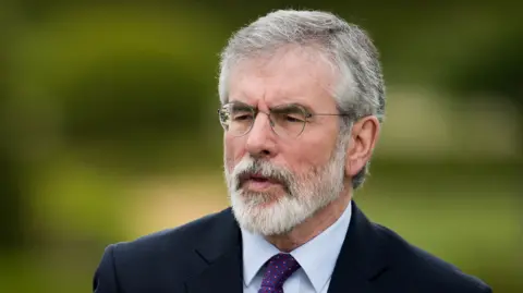 Getty Images Gerry Adams looks into the camera. He is wearing round glasses, a navy jacket, white shirt and black and gold patterned tie. He has white hair and a white beard.