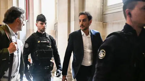 Getty Images Defendant Gary Madar walking in the courthouse in Paris, with a police officer walking in front of him and another behind him