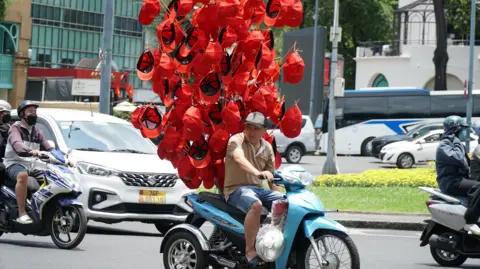 BBC/ Lulu Luo A street seller on a motorbike carrying scores of red caps to sell.