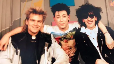 Mike Peters, Gaz Top and a friend pose for a photo on the Music Box channel. Their hairstyles are classic 1980's style as well as the outfits