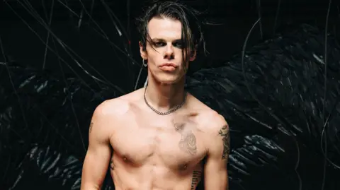 BBC Yungblud poses against a backdrop of black feathered fans, with his shirt off and multiple tattoos visible.