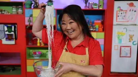 ABC Kids A woman in a red shirt and yellow apron lifting noodles out of a mixing bowl with a pair of tongs