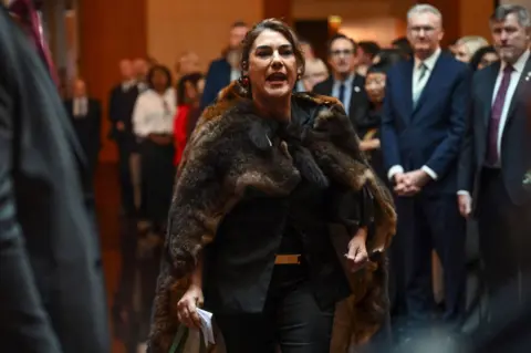 Getty Images Lidia Thorpe wearing a fur coat holds a piece of paper while standing in a room in front of men in suits