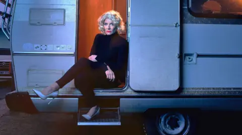Michael Wharley The actress Michelle Collins in a Marilyn Monroe styled blonde curly wig and black polo neck and leggings with white stilleto shoes sits on the steps of a rather grubby caravan. She looks very angry.