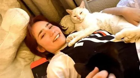 Family handout Elise Sebastian lies on cushions with curtains in the background, she is smiling and looking at the camera and has a white cat on her lap. She has her arm around the cat. Elise has brown hair and is wearing and black and white top