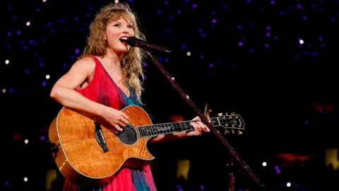 Getty Images Taylor Swift performing on stage during her Eras tour. She has her long blonde hair curled and wears a red and green flowy dress while holding an acoustic guitar and singing into a mic on a stand.