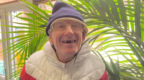 A man wearing glasses and a white puffer jacket, smiling in front of a large plant.