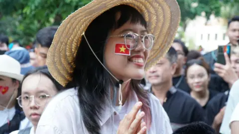 BBC/ Lulu Luo Tung Linh smiles in a hat and with the red and yellow Vietnamese on her cheek. Behind her people are wlaking on the street