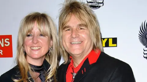 Getty Images Jules and Mike peters. They both have long blonde hair, are wearing black jackets, and are smiling at the camera