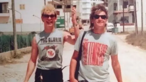 Gaz Top and Mike Peters walking down a street in Ibiza, Spain. There is a road behidn them lined with buildings. Gaz is wearing a The Alarm top. Mike is wearing a t-shirt with some red writing on it