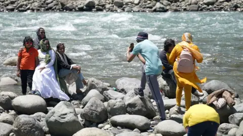 Seraj Ali The image shows tourists sitting on the rocks by a river in Pahalgam town, with some families posing for a photograph.