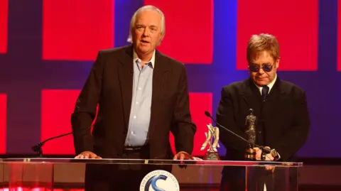 Sir Tim Rice stands next to Sir Elton John at the Ivor Novello Awards podium