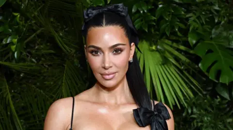 Getty Images Kim Kardashian looks straight down the camera with her hair in a high pony tail and lots of palm tree leaves behind her.