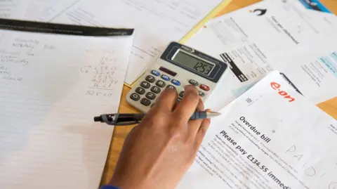 Getty Images A close up of a woman's hand as she calculates her energy bill and other outgoings using a calculator and a lined pad of A4 paper - her right hand is holding a pen and pressing numbers on the calculator and bills are spread over the table