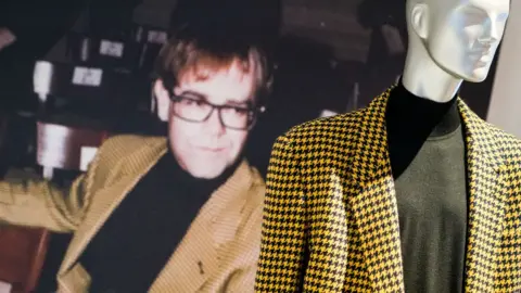 Versace Retrospective A yellow blazer worn by Sir Elton John, shown as part of the exhibition and featured next to a picture of the singer wearing it