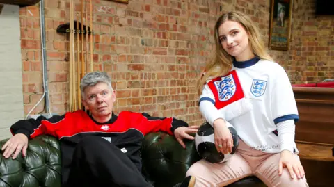 @thoughtsofawing Christine Lockwood, sitting on a green sofa, with her leg across her other leg, looking at the camera. She is wearing black trousers, trainers and a black and red top. Nicole Lockwood-West is sat on a stool to her right, wearing a football top, pink trousers, trainers, and is holding a football. A pool table is behind them, showing a brick wall.