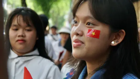 BBC/ Lulu Luo Minh in a blue denim shirt with a flag sticker on her cheek, wearing two earrings in her ear. Another girl in a white top stands to the side