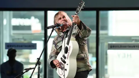 Nicholas Hunt/Getty Images Mike Peters who has long blonde hair and is wearing a khaki camouflage jacket. He is stood at a microphone stand and is holding a cream guitar which he is playing and looking at. Behind him are some large windows and a man wearing a baseball cap.
