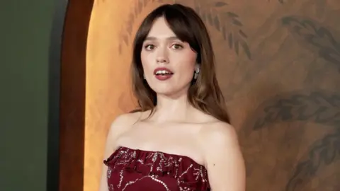 Getty Images Aimee Lou Wood, who has long brown hair and is wearing a strapless burgundy dress, poses at a The White Lotus premiere in Los Angeles