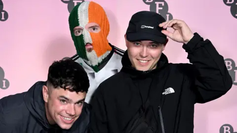 Getty Images Three young men are standing close to each other posing for a photo. They are standing in front of a pink backdrop. One is crouched down and smiling. The man in the middle is wearing a Irish tricolour balaclava.