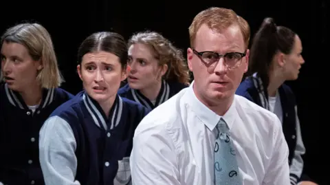 Lloyd McDonagh A still from a play, showing a man in the middle, wearing a white shirt and tie, with glasses on and short ginger hair. Four women are behind him, wearing blue jackets, looking shocked.