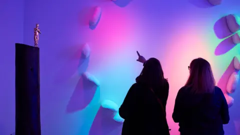 Stay a While, Stuart Calvin Women pointing at sculptures of rocks placed in a circle with added colourful lighting at the Golden Thread Gallery in Belfast