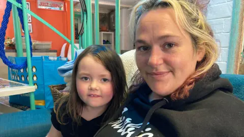 A woman wearing a black hoodie, sat on a sofa with a young child. The young child wears a black top with long dark hair. Her mum has shorter, blonde hair.