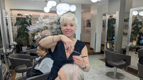 BBC Kerry Larcher cuts a customer's hair in her salon and smiles for the camera