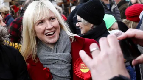 Reuters MP Rosie Duffield, wearing a red coat, grey scarf and a red Labour rosette, smiling as she is being photographed in a press huddle.