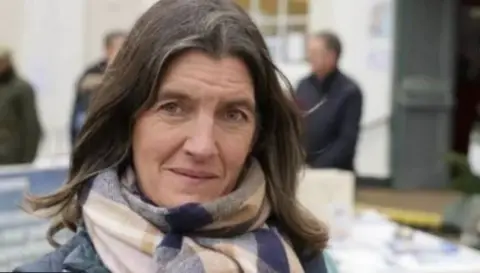 John Fairhall/BBC Rosie Pearson with mid-length brown hair looks into the camera. She is wearing a scarf with a purple, beige and pink checked pattern.