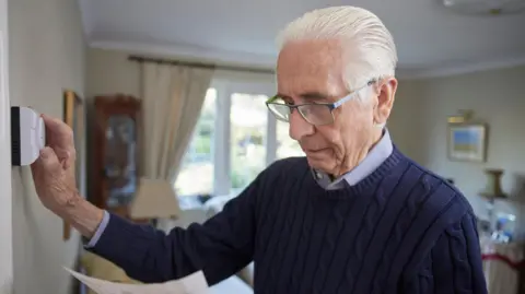 Getty Images An elderly gentleman wears a blue jumper in his living room at home. He has an energy bill in one hand and is adjusting the thermostat with the other.