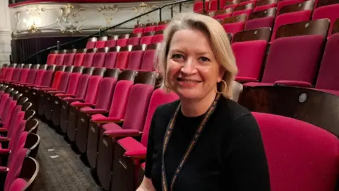 BBC/Seb Cheer A woman with blonde hair sits in an empty theatre auditorium. She is looking at the camera and smiling. Her lanyard says