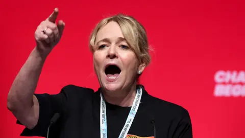 Reuters A woman with blonde hair tied back in a ponytail stands in front of a red background. She is wearing a black top and has a lanyard round her neck. She is gesticulating with her hand raised and forefinger pointing and she appears to be shouting or at least talking animatedly.
