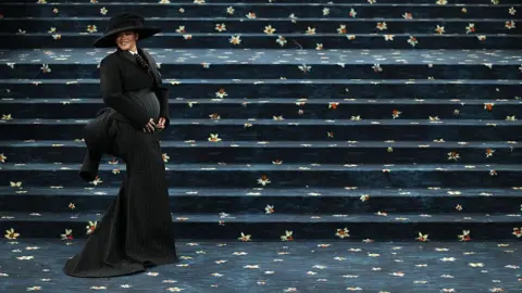 Getty Images Rihanna smiles and cradles her baby bump as she appears on the red carpet, which is actually navy with daffodils, for the 2025 met Gala. She is wearing a wide-brimmed hat and a floor-length tailored outfit.