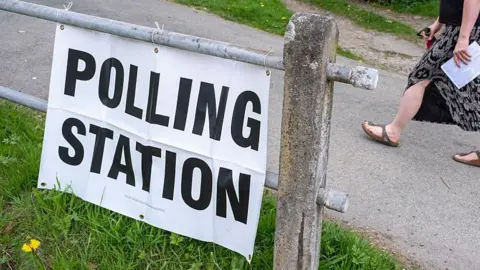 Getty Images A polling station in Shropshire. There is a metal gate with a paper sign that says POLLING STATION. Someone can be seen walking past with a ballot paper in the background