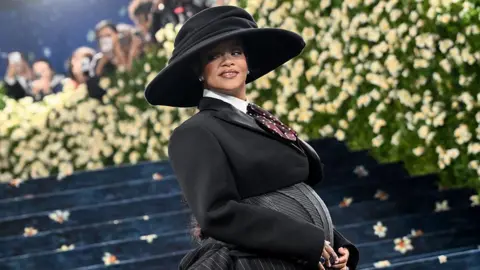 Getty Images Rihanna appears at the 2025 Met Gala visibly pregnant. She is wearing a black and grey tailored outfit and large brim hat.