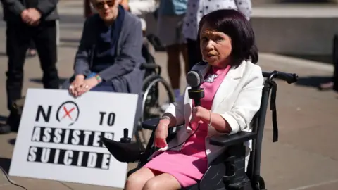 PA Media A woman with dark hair, wearing a pink dress and white jacket, speaks into a microphone outside the Scottish Parliament. She is sitting in a black wheelchair. A person behind her is holding a