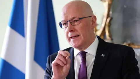 Getty Images John Swinney, a blad man waring glasses, a dark suit with white shirt and purple tie, is standing in front of a Saltire flag with his right hand raised as he answers a question from an unseen group