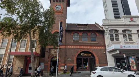 BBC/Kelly Ng People walk along the pavement in front of a clock tower at Burwood, Reid, next to a Bank of China branch office