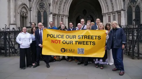 PA Media A group of people standing outside the Royal Courts of Justice holding a yellow banner with the slogan