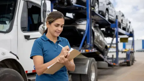 Getty Images A woman with long straight brown hair in a ponytail wearing a blue uniform style polo shirt writing on a clipboard in front of a lorry full of cars